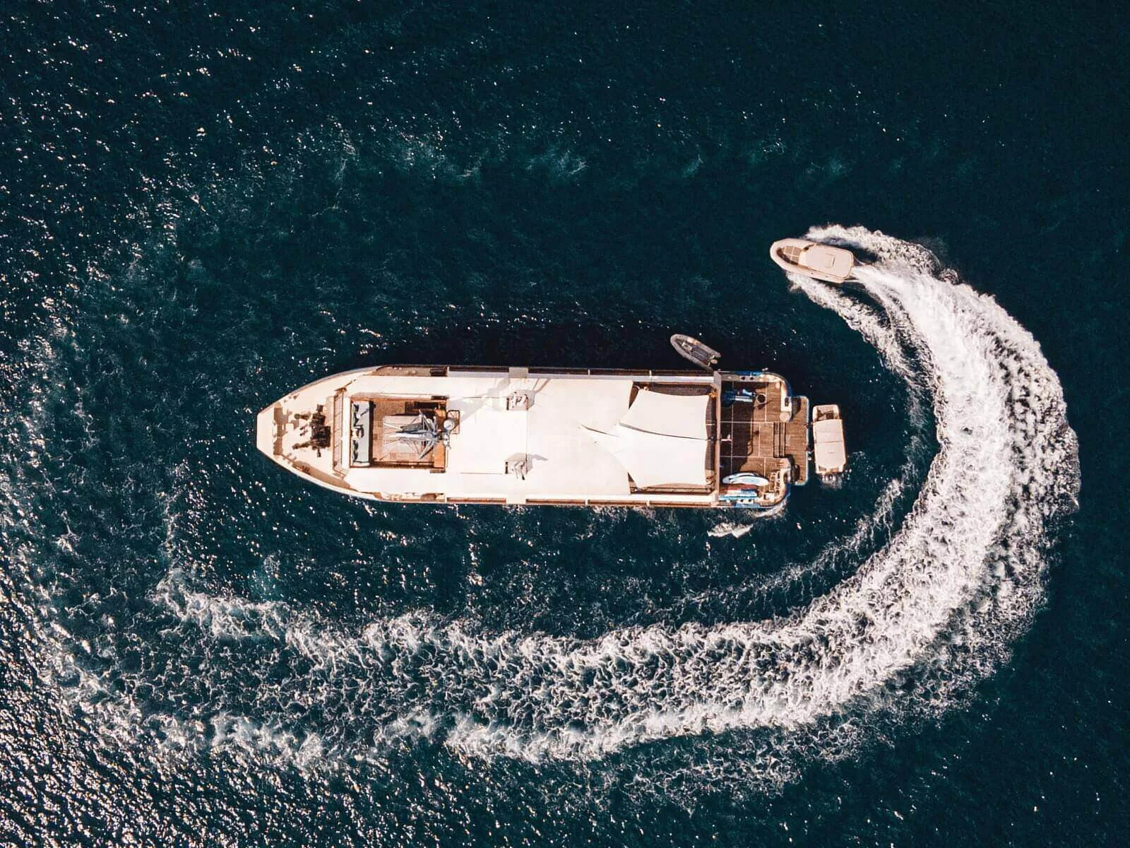 Aerial of the Kudanil Explorer expedition vessel with a tender carving a curved wake on dark water